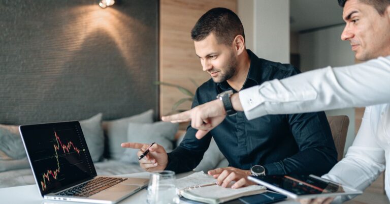 Como Usar Candlesticks no Trading de Forex: Um Guia para Traders Iniciantes Dois homens discutindo dados financeiros em um laptop em um ambiente de trabalho moderno